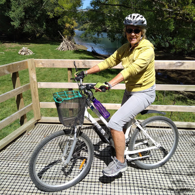 Woman riding a bicycle on a wooden platform with a scenic background of trees and water.